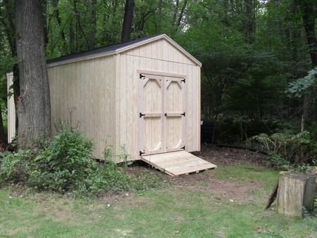 Wooden shed with ramp in a wooded area, surrounded by trees and bushes.