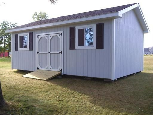 Light blue shed with white trim, double doors, two windows with brown shutters, and a wooden ramp on a grassy lawn.