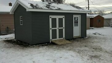 Gray shed with double doors and a ramp on a snowy day.