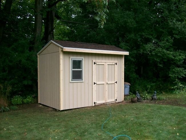 Tan shed with a dark roof and a window. It has double doors in a grassy backyard.