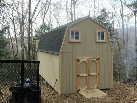 Tan barn with black roof, two windows, and double doors, set in a wooded area.