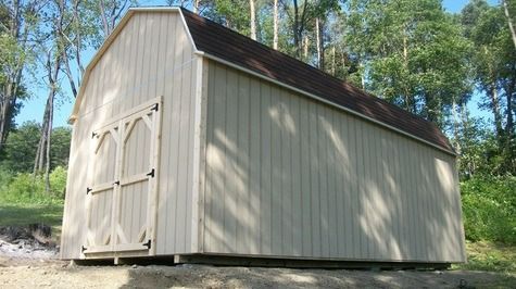 Tan barn-shaped shed with brown roof and double doors in a grassy, wooded area.
