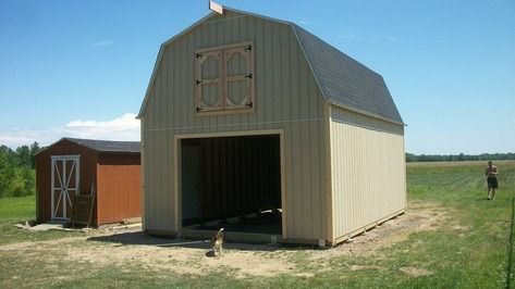 Tan barn with open front and a smaller red shed. Person standing in a grassy field.