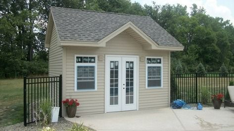 Small tan shed with a gabled roof, white-framed windows, and French doors, surrounded by a black fence.