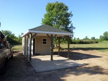 Small building with a covered porch on a concrete pad, set in a grassy outdoor area.