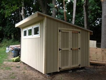 Tan shed with windows and double doors, built in a wooded backyard.