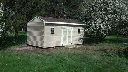 Tan shed with brown roof, white doors and small windows, set in a grassy yard.