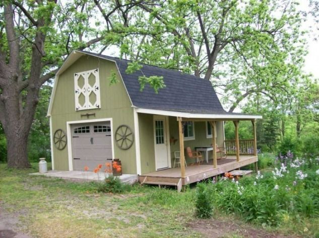 Green barn with a porch, garage door, and wagon wheel decorations in a grassy yard.