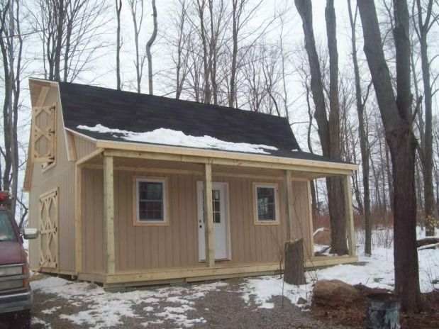 Small tan cabin with a porch and black roof in a snowy wooded area.