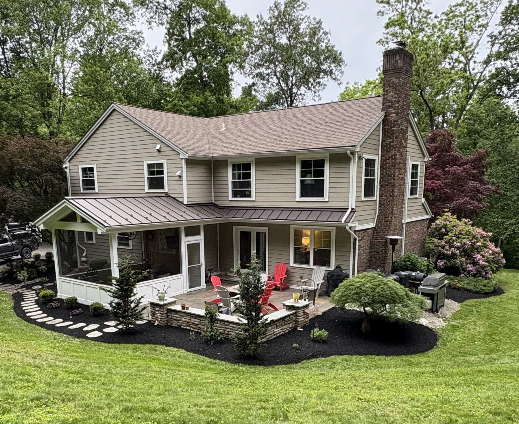 Two-story house with backyard patio, landscaped beds, and chimney, surrounded by trees.