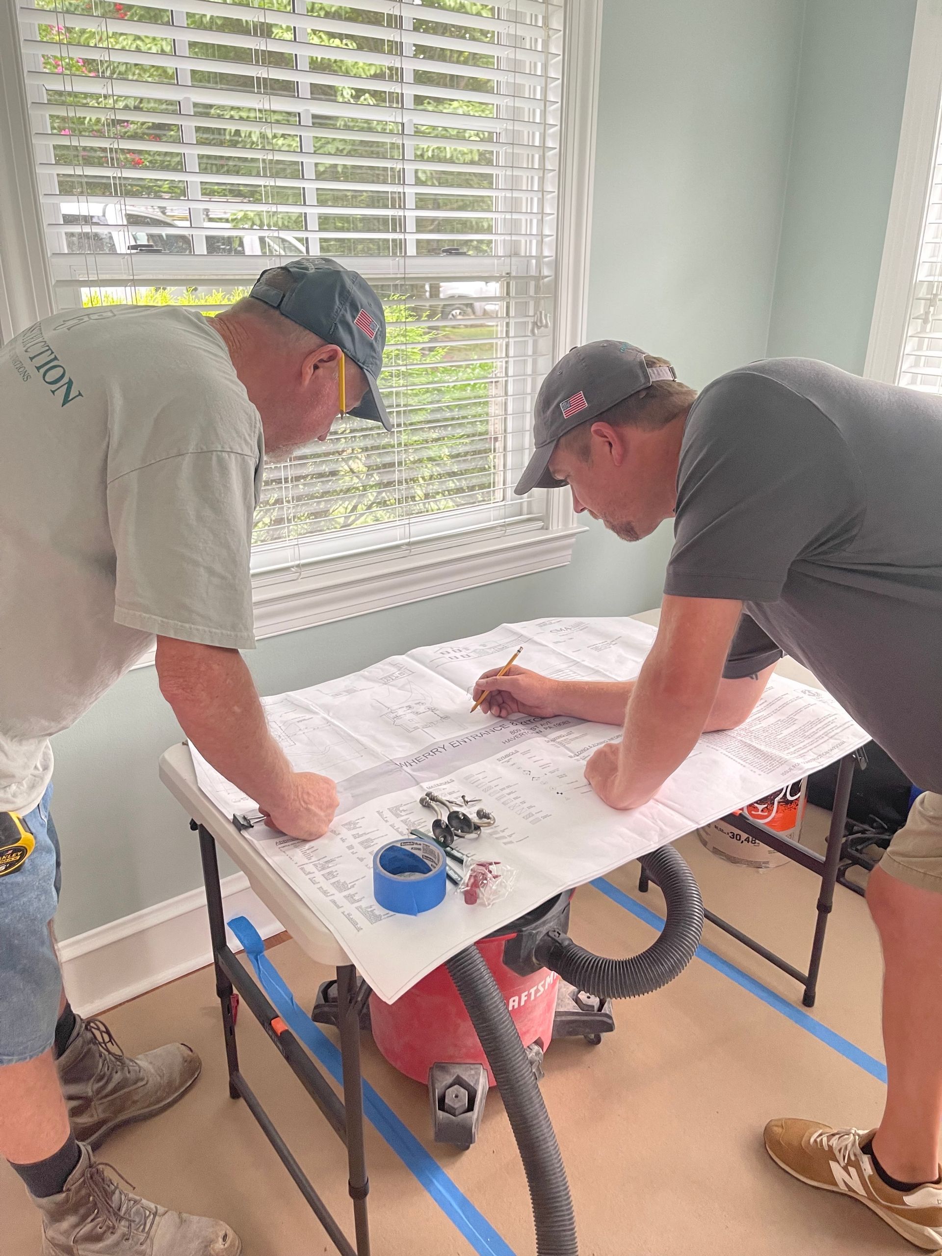 Two men in hats looking at blueprints at a table near a window.