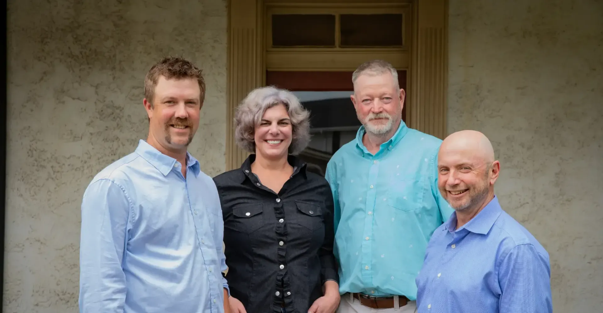 Four people standing outside a building: two men on the ends, two in the middle. All are smiling.