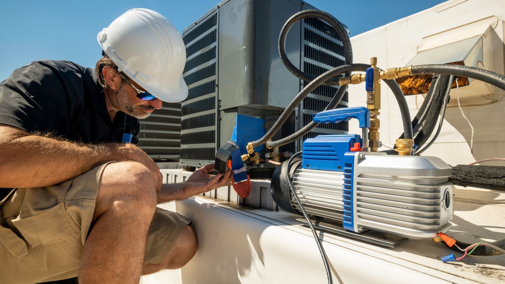 HVAC technician repairs rooftop unit, inspecting connections with vacuum pump. Sunny day, blue pump, tan shorts.