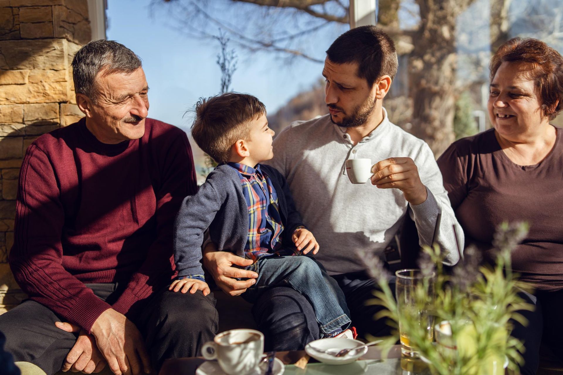 Family, child on father's lap, seated outdoors, having coffee. Smiling, sunny.
