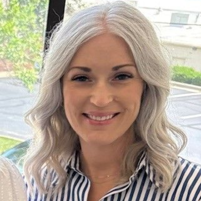 Blonde woman smiling, wearing a light blue sweater and necklace, sitting outdoors in autumn.