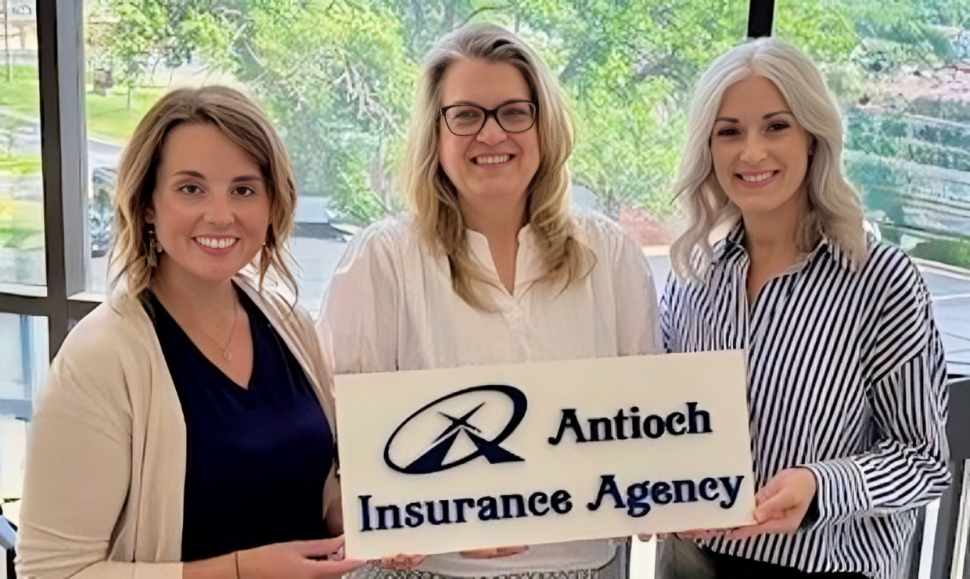 Three women hold an Antioch Insurance Agency sign, smiling inside by a window.