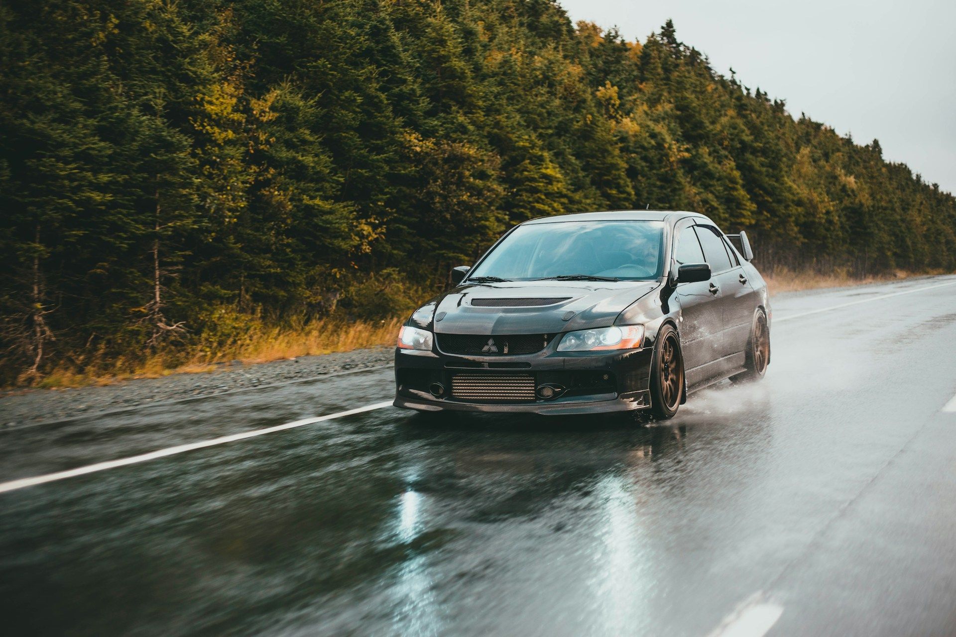 Black Mitsubishi Lancer Evolution driving on a wet road, with trees in the background.