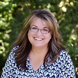 Woman with glasses smiles; she's wearing a patterned shirt with greenery in the background.