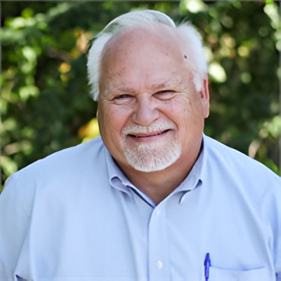 Smiling older white man with white hair and goatee, wearing a light blue shirt, outdoors.