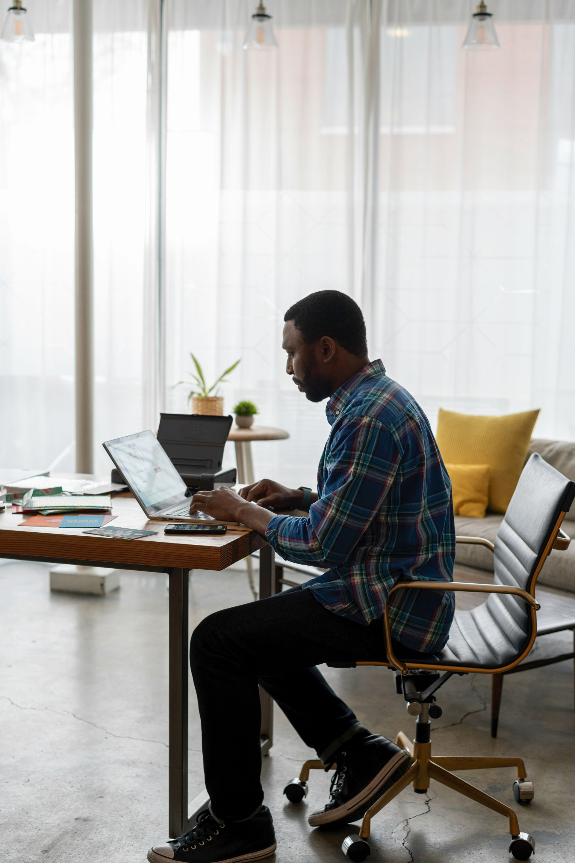 Man in blue plaid shirt works on laptop at a desk in a modern office, side view.