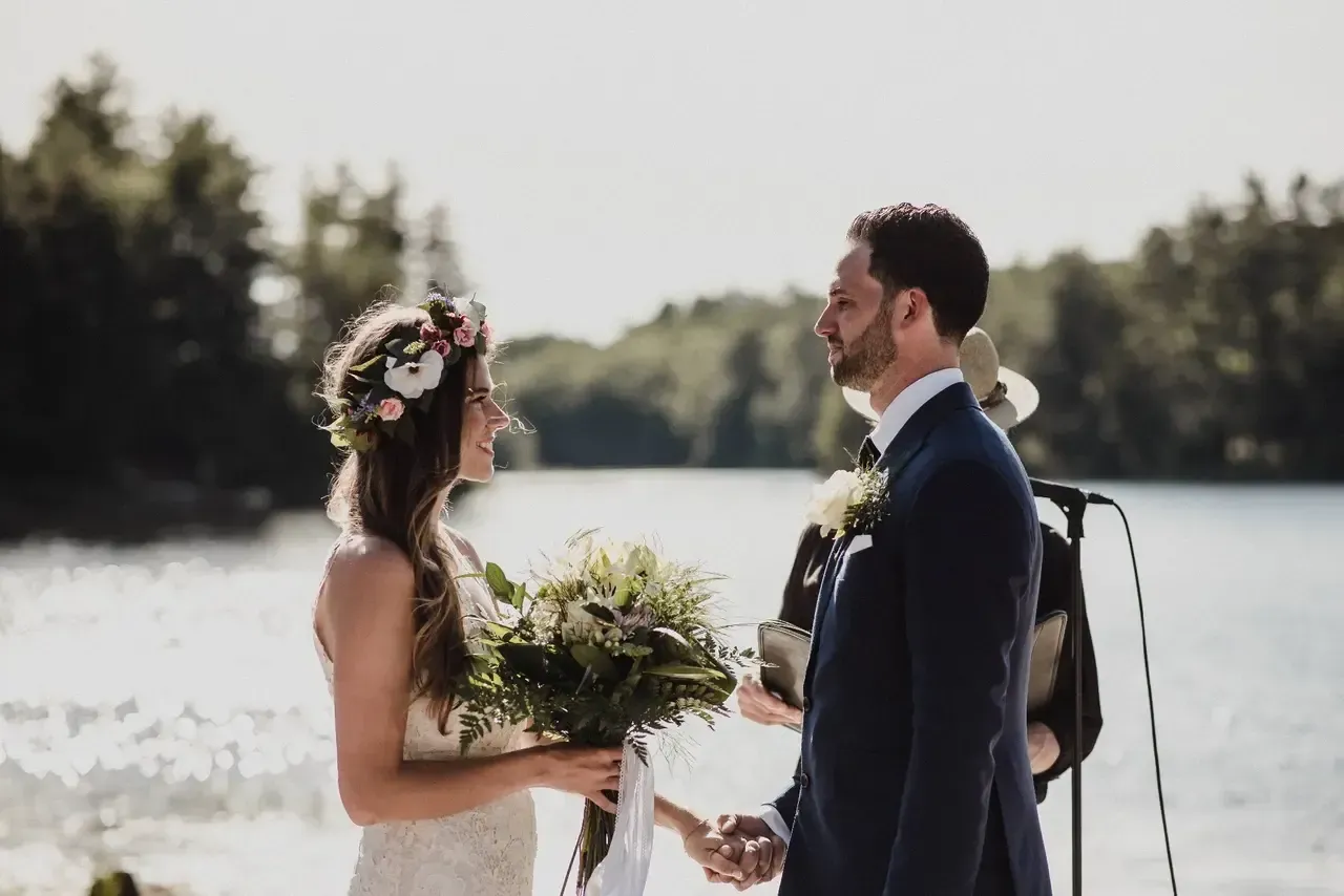 A bride and groom are holding hands during their wedding ceremony in front of a lake.