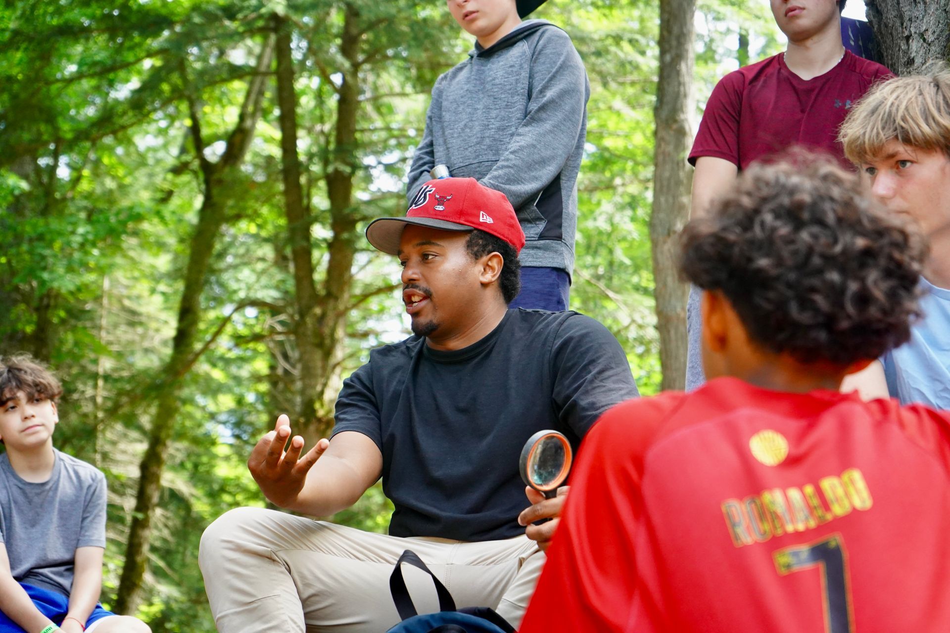 A man is sitting in the woods talking to a group of young boys.