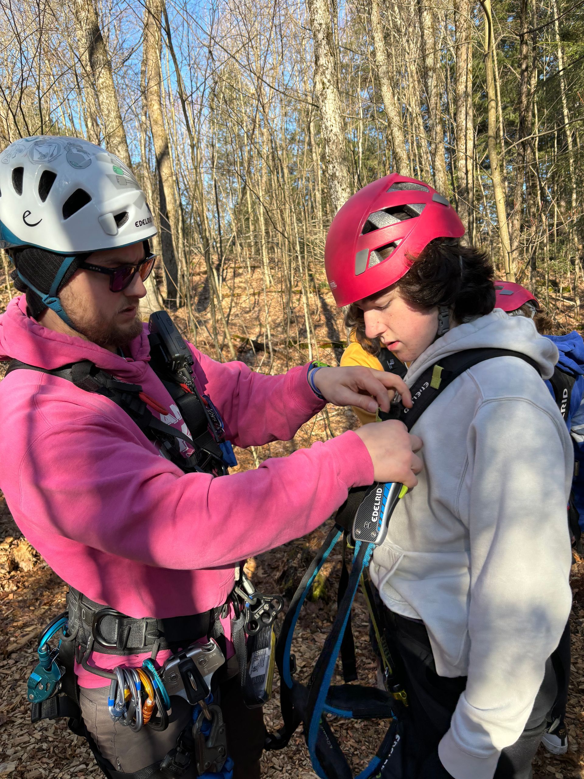 A Camp Muskoka staff member  is helping a male student put on a helmet in the woods.