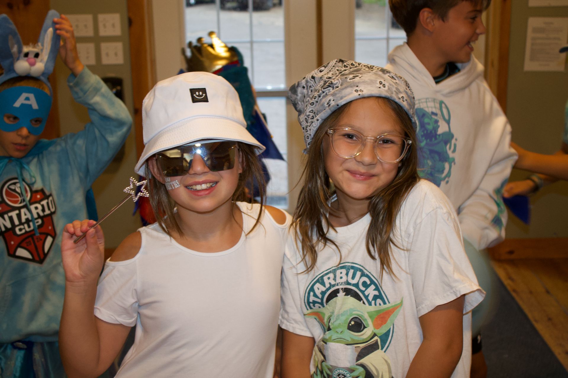 Two girls wearing sunglasses and hats smile, posing for a photo. A boy in a costume is behind them.