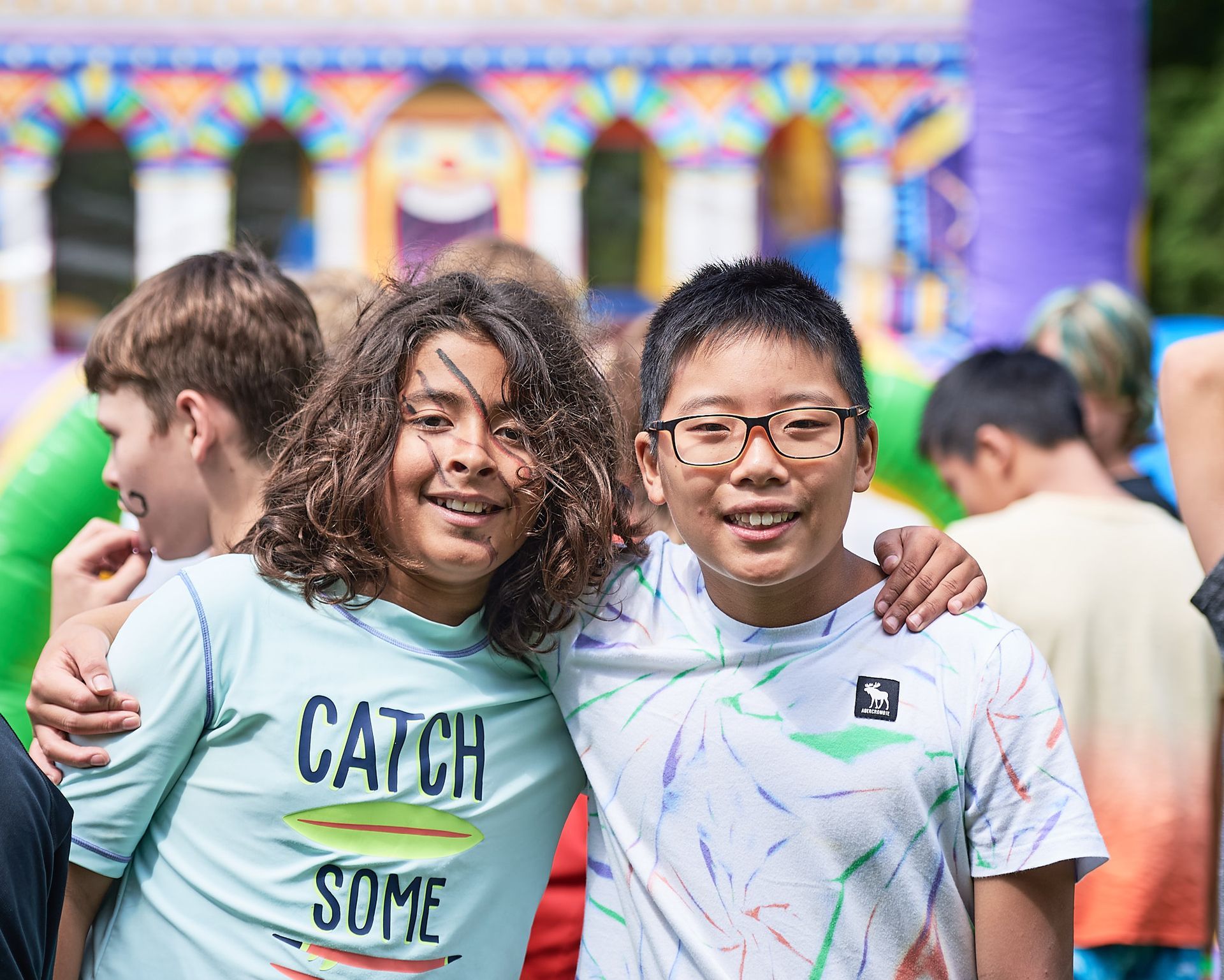 Two smiling boys with arms around each other pose for a photo at an outdoor event with a colorful bounce house in the background.