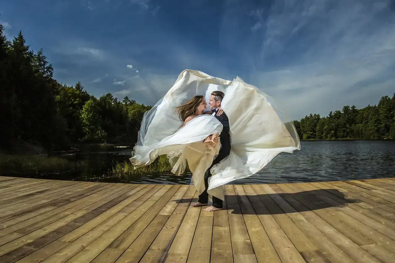 A bride and groom are dancing on a dock near a lake.