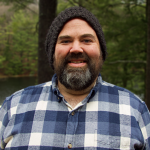 A man with a beard is wearing a plaid shirt and a hat.