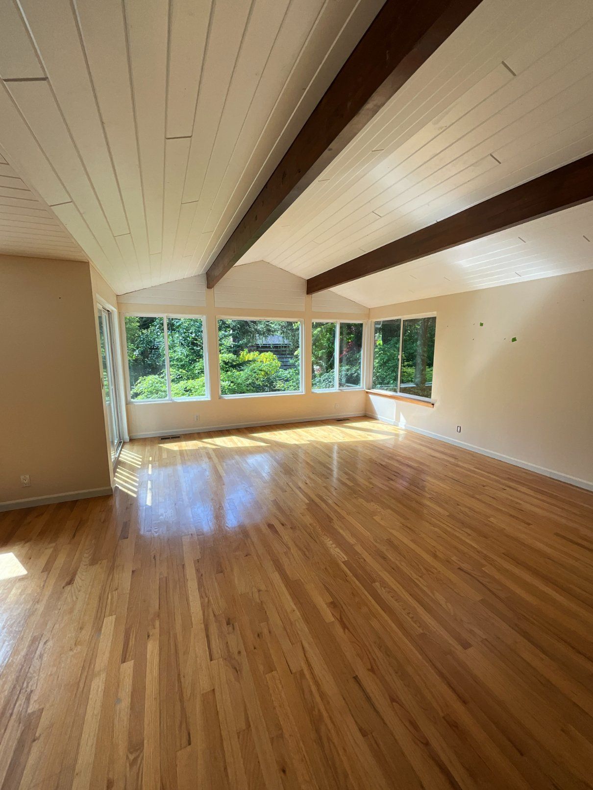Spacious room with wooden floor, large windows, and a sloped, wood-beamed ceiling. Sunlight streams in.
