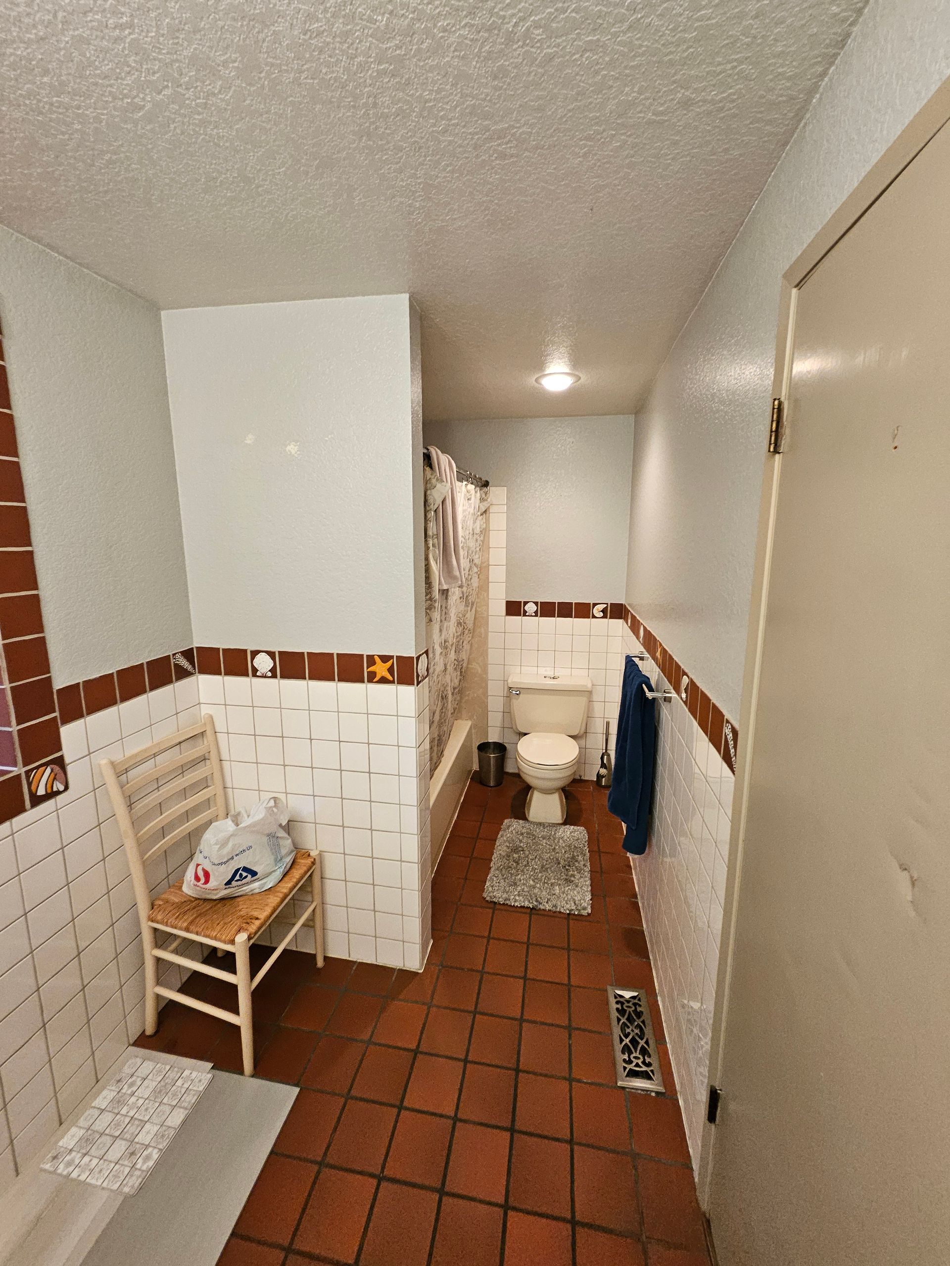 Bathroom with white and brown tiles, toilet, chair, and a shower curtain.