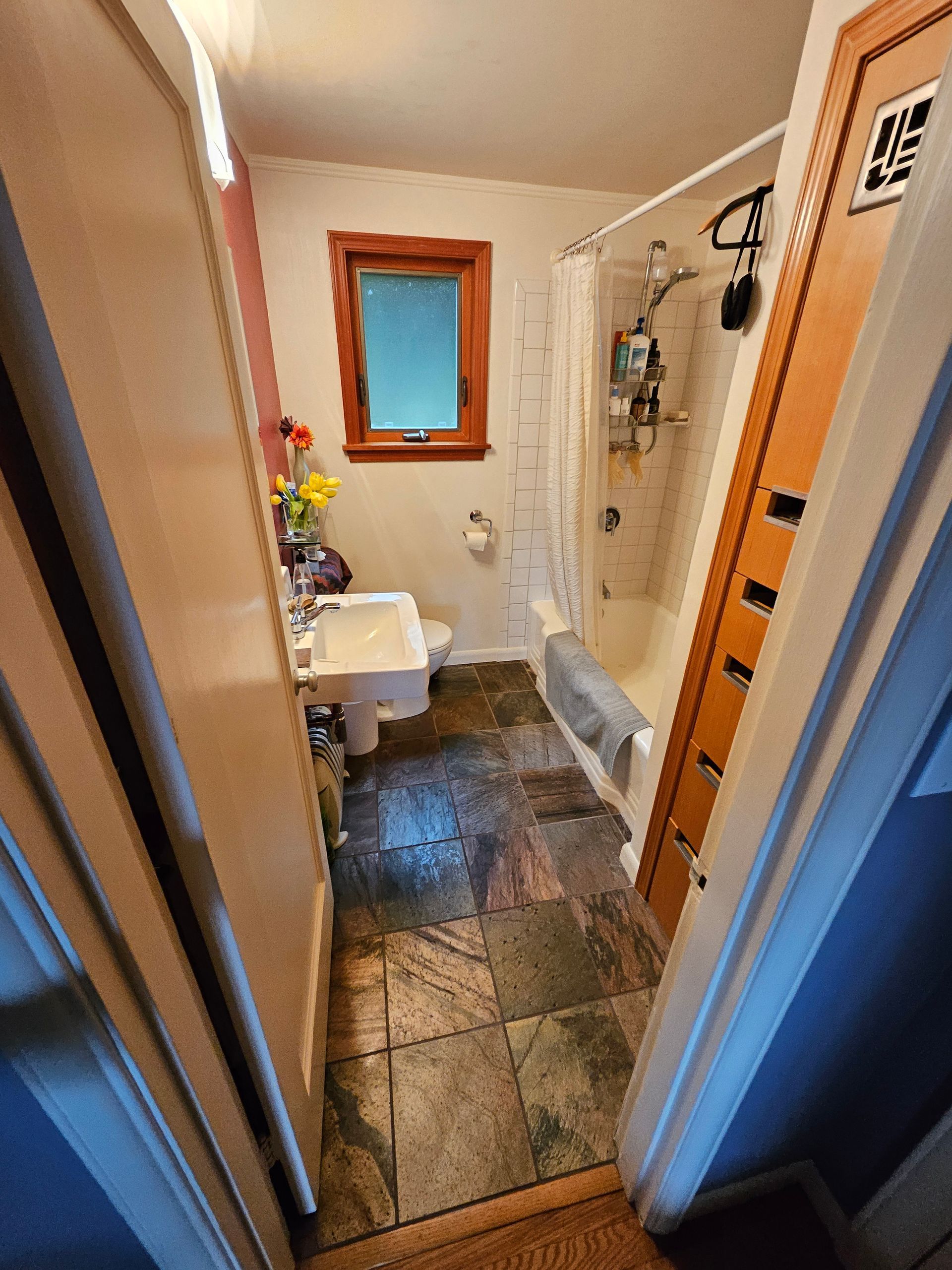 Bathroom with slate tile floor, shower, toilet, sink, and window with wooden frame.