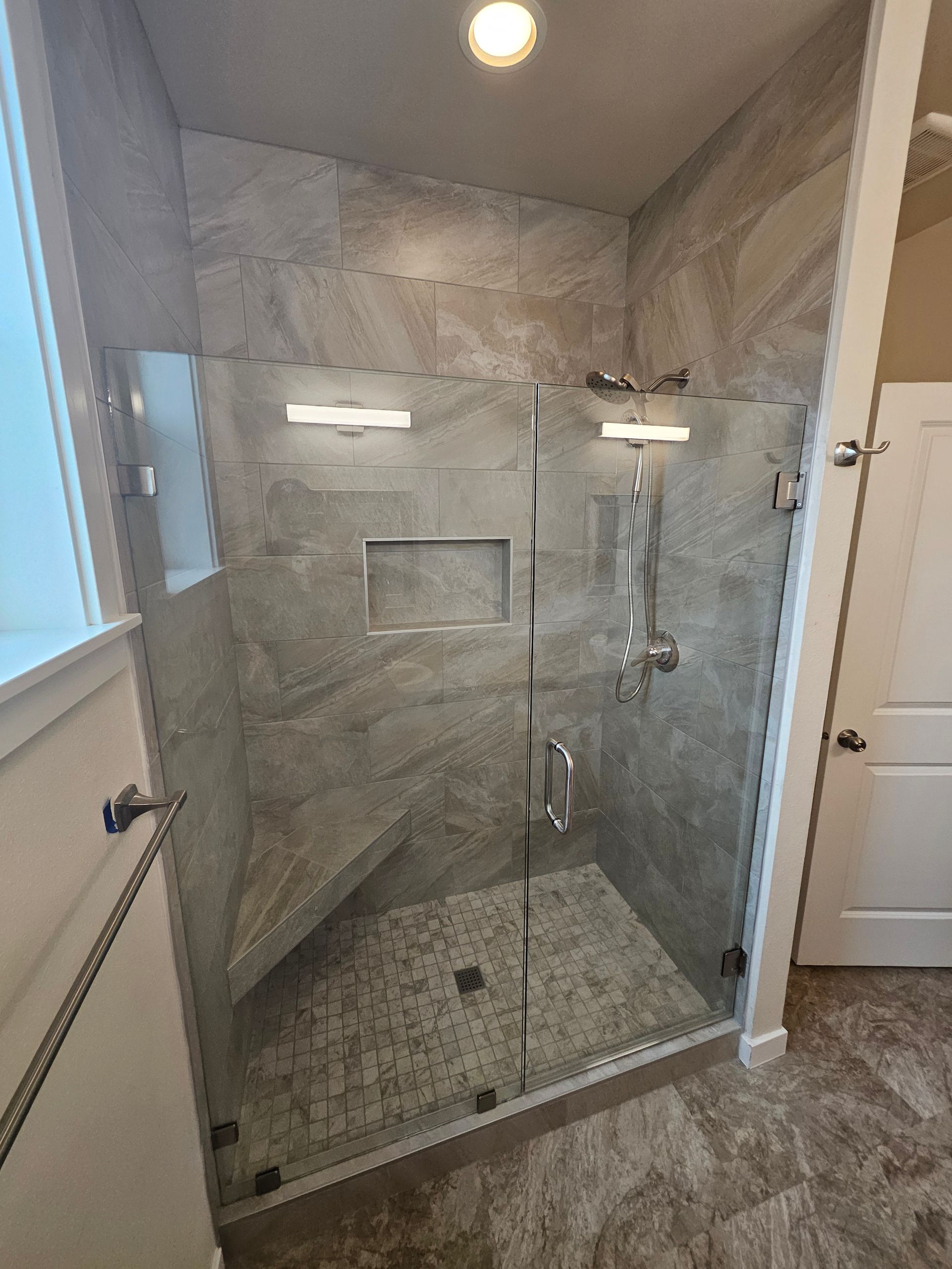 Modern walk-in shower with gray tile, glass doors, built-in bench, and rain showerhead.