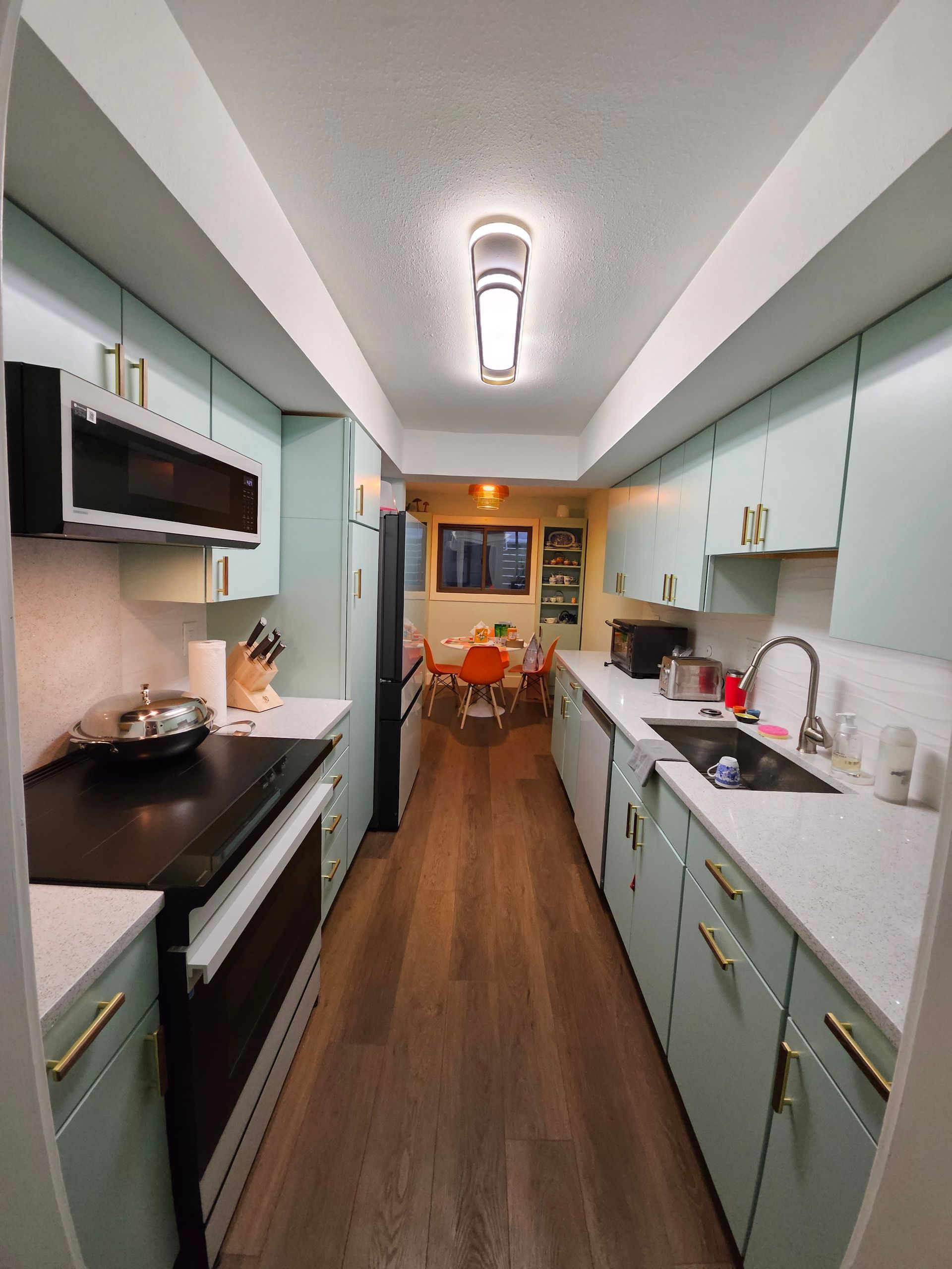 Narrow kitchen with light blue cabinets, dark oven, and wood floor. Open to dining area.