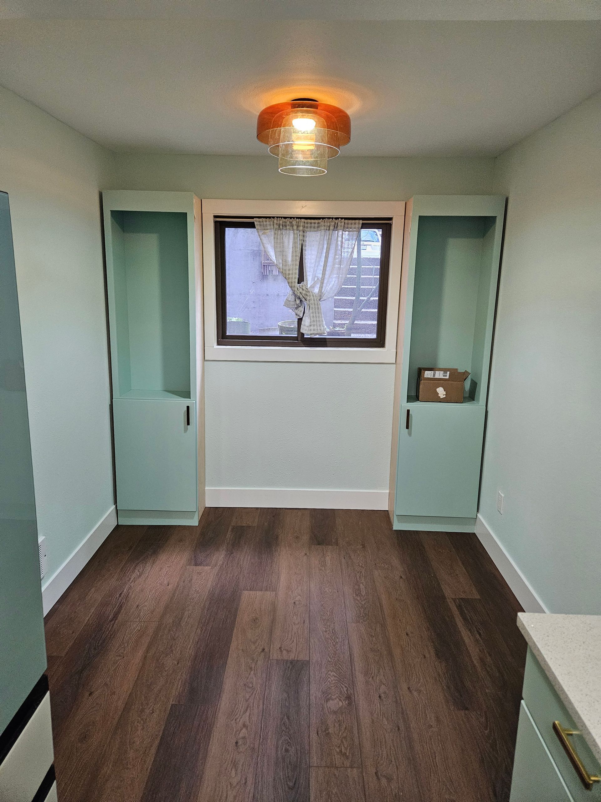 Small room with built-in teal storage cabinets on either side of a window. Brown wood floor.