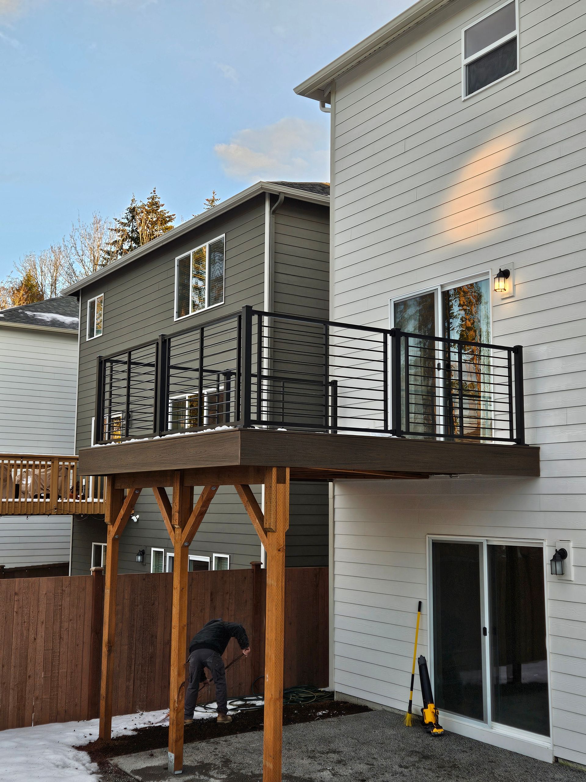Two-story house with a wooden deck supported by wooden posts. A person is working in the yard.