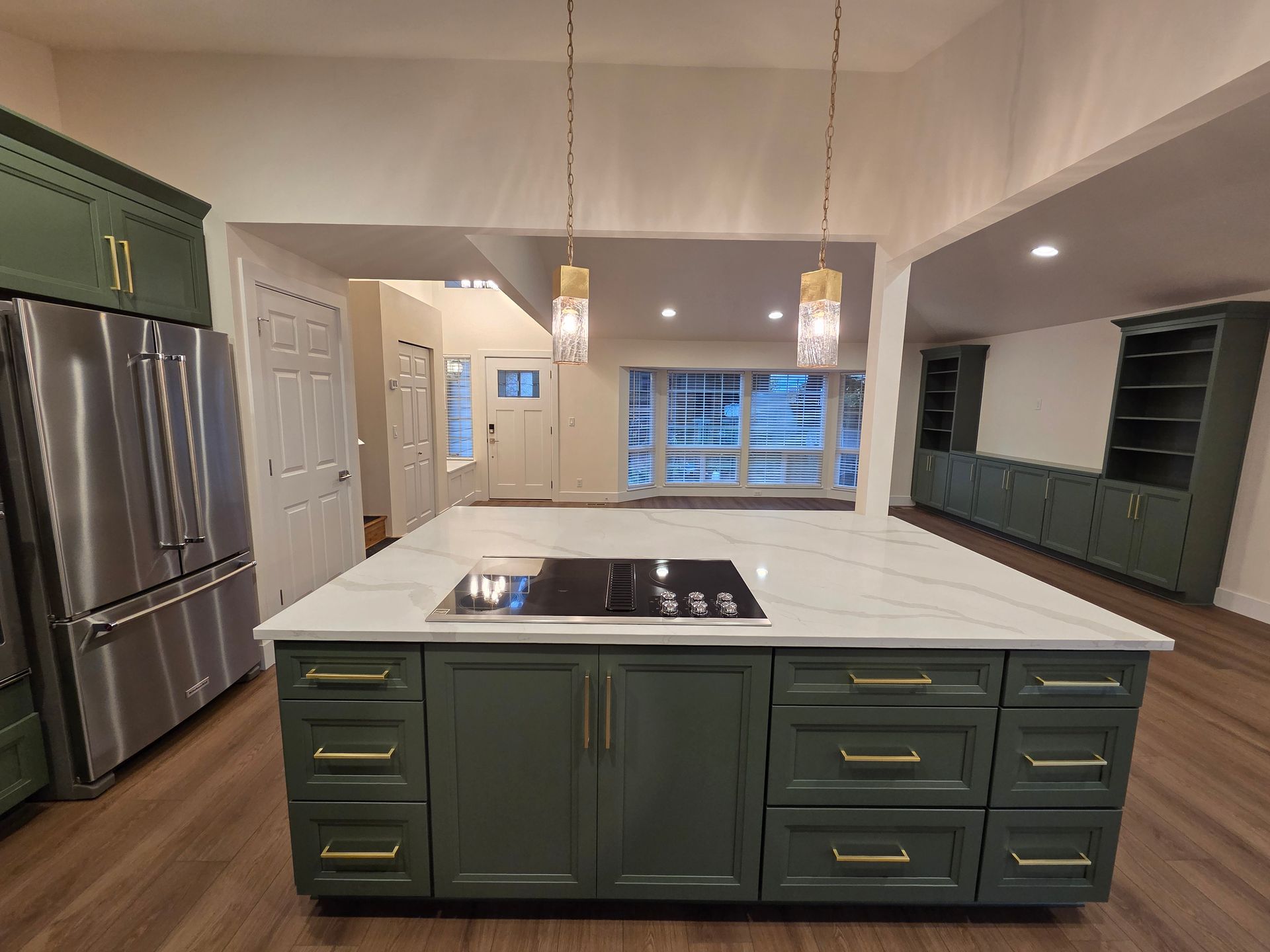 Green kitchen with island and white countertop, stainless steel refrigerator, and hanging lights.