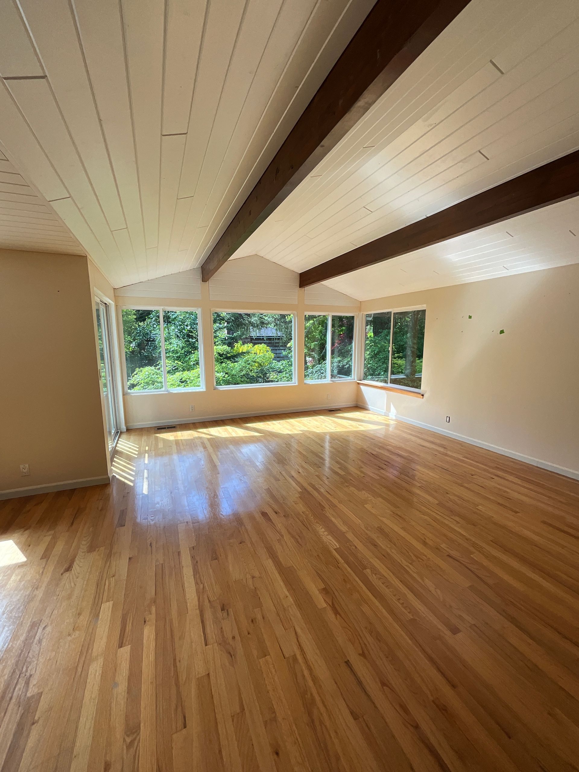 Empty room with hardwood floors, large windows, and exposed wooden beams.
