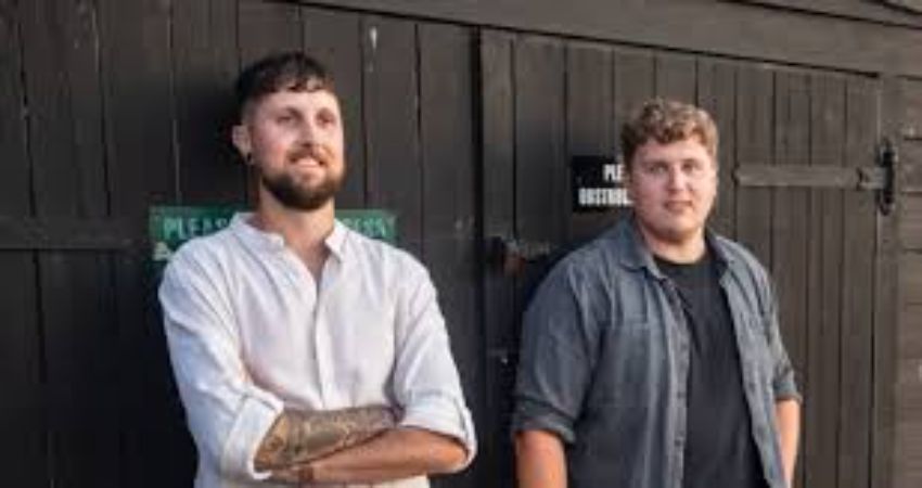 Two men are standing next to each other in front of a wooden building.