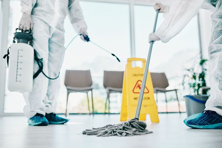 Two people in hazmat suits cleaning an interior. One sprays, the other mops. A wet floor sign is present.