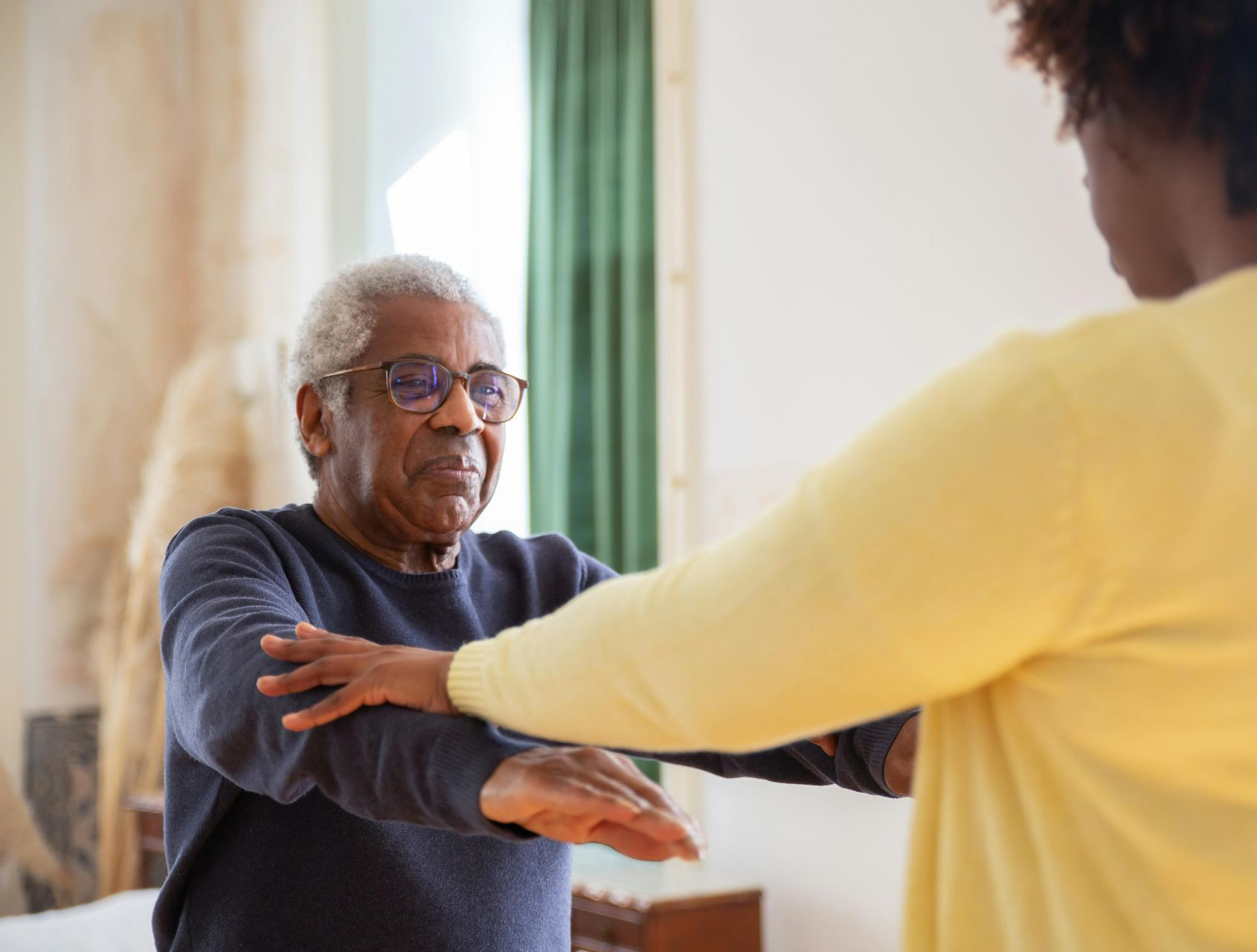 Older Black man doing arm exercises with the help of a young woman in a room.