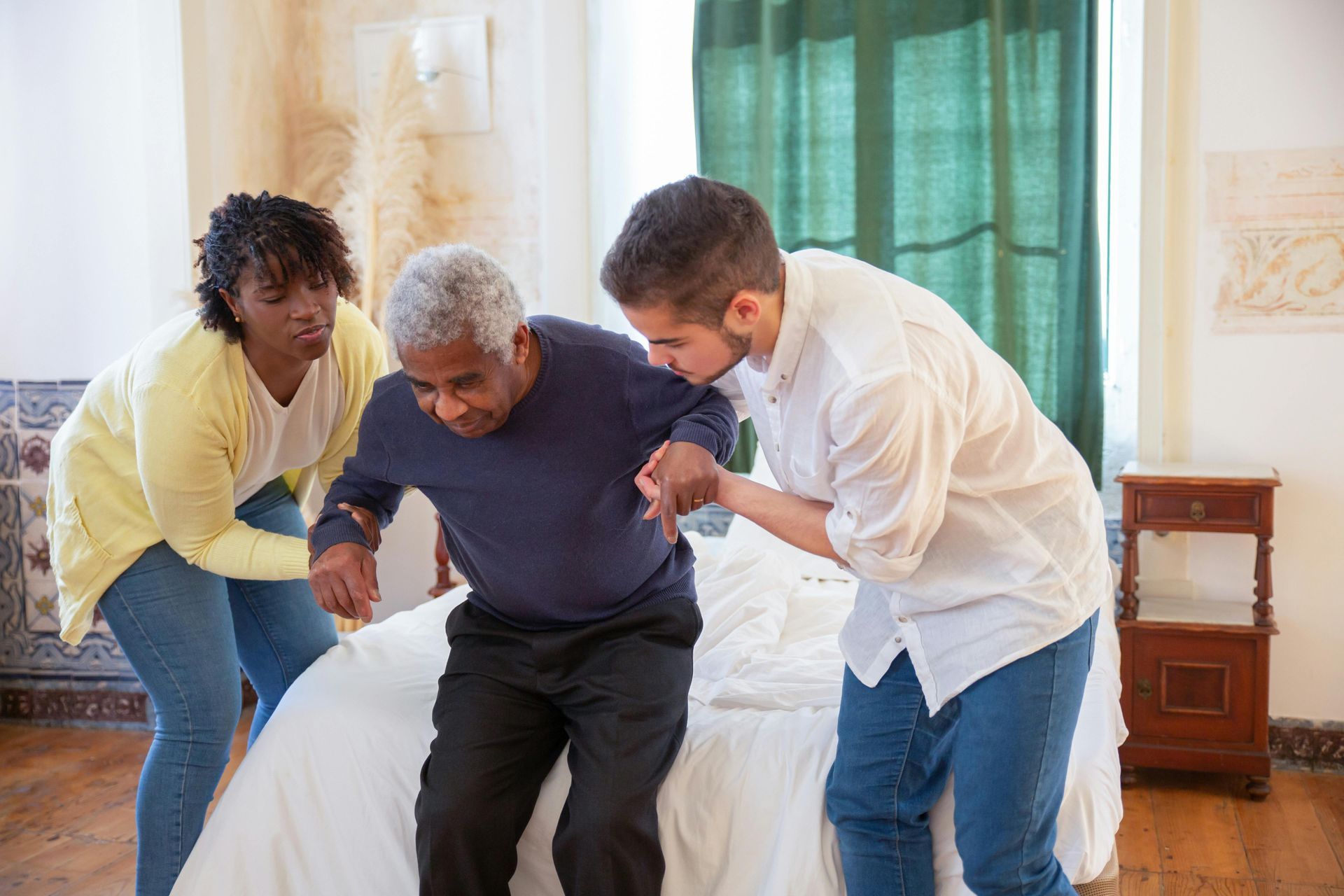 Two people assisting an elderly Black man getting out of bed. They are in a bedroom.