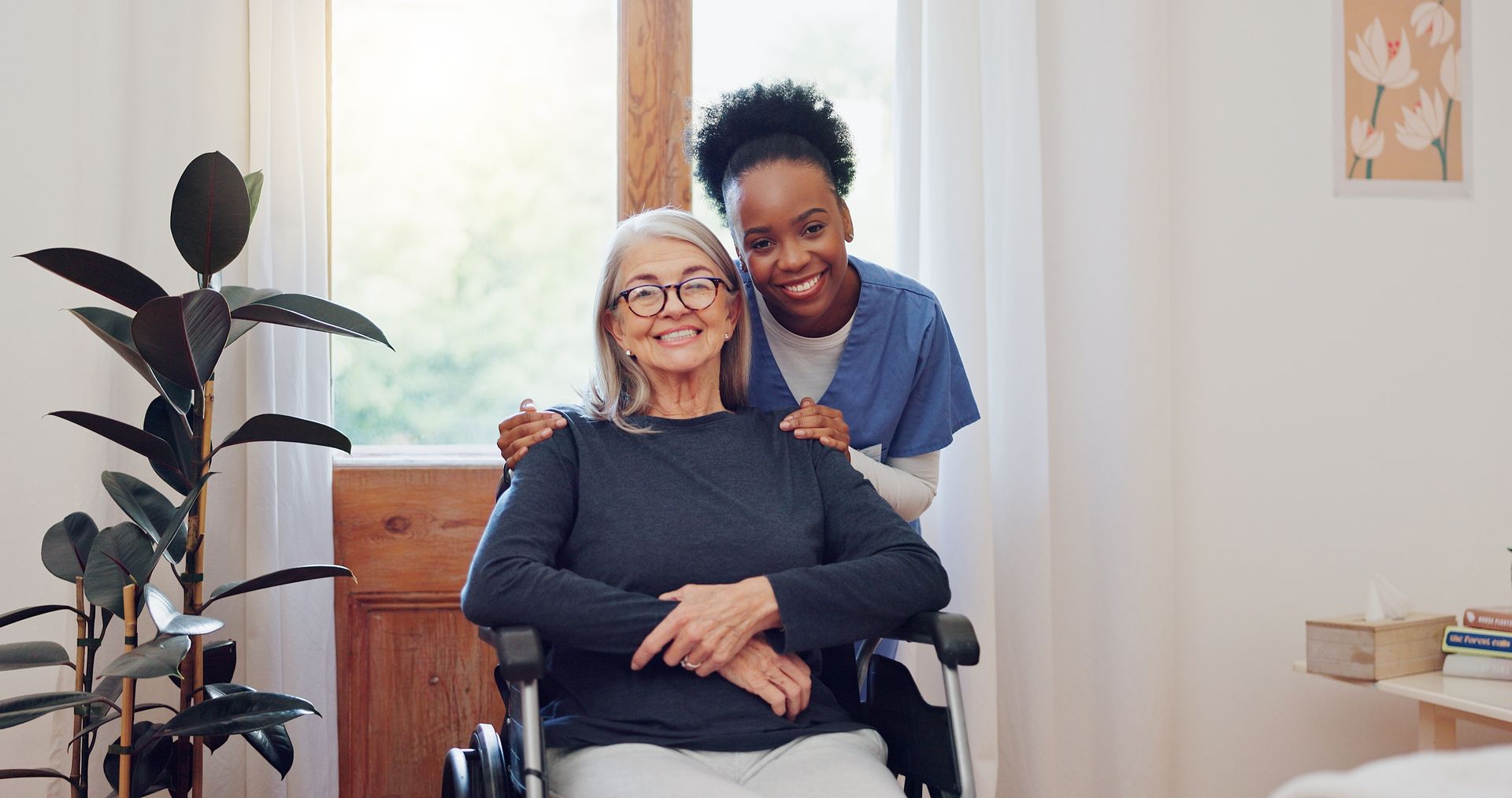 A senior woman in a wheelchair smiles with a healthcare worker, indoors near a window.