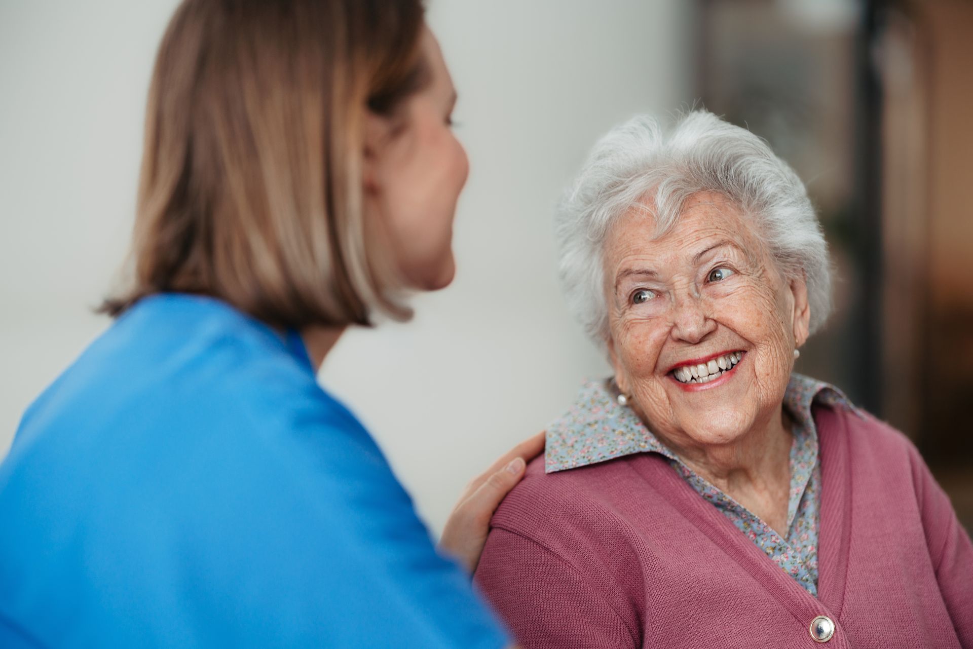 Nurse in blue scrubs smiles at an elderly woman wearing pink cardigan. Inside, soft lighting.