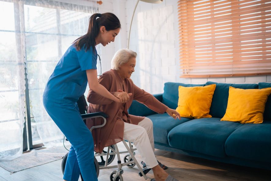 Caregiver in blue scrubs helps elderly woman from wheelchair to sofa.