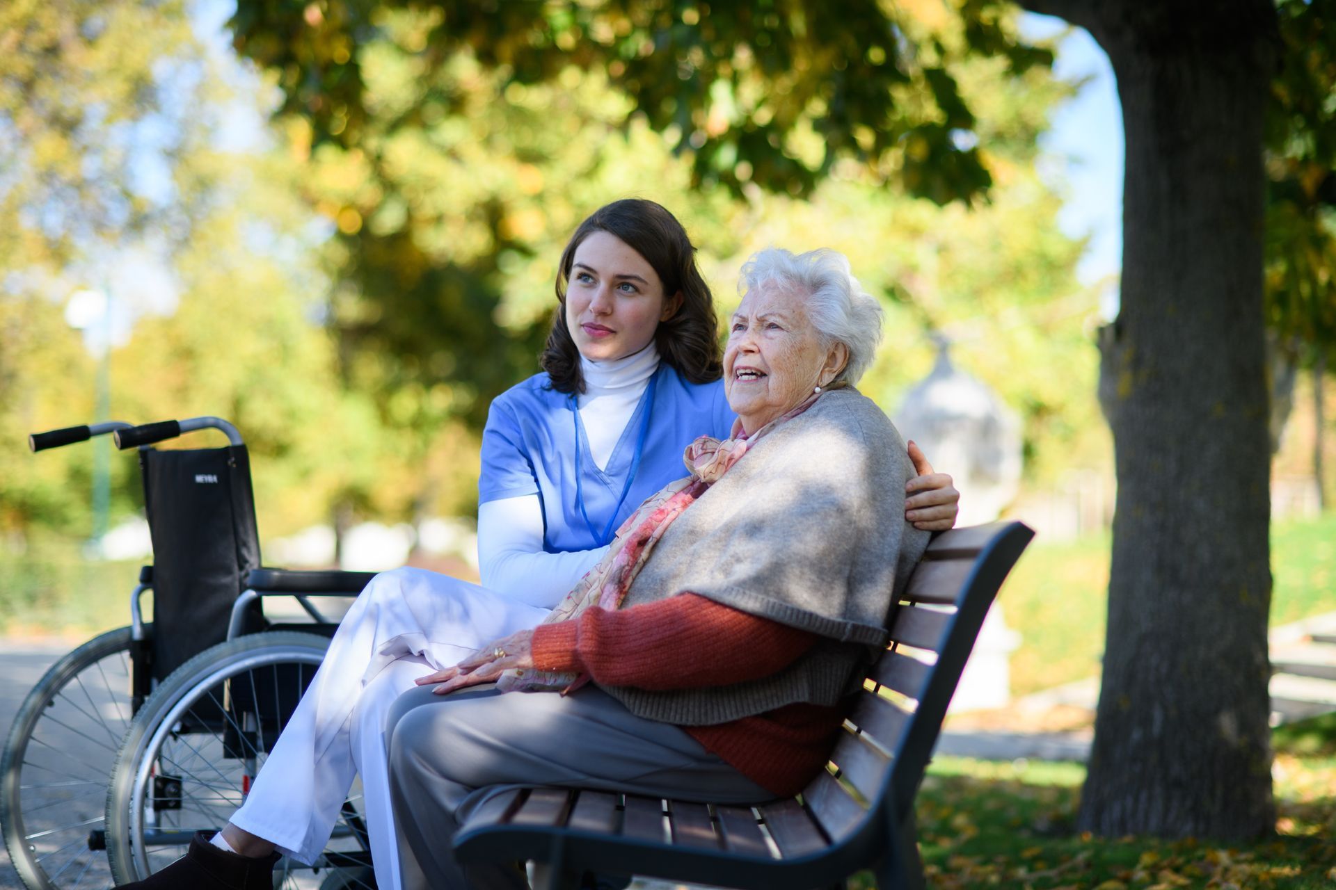 Young woman in scrubs comforts elderly woman on park bench next to a wheelchair.