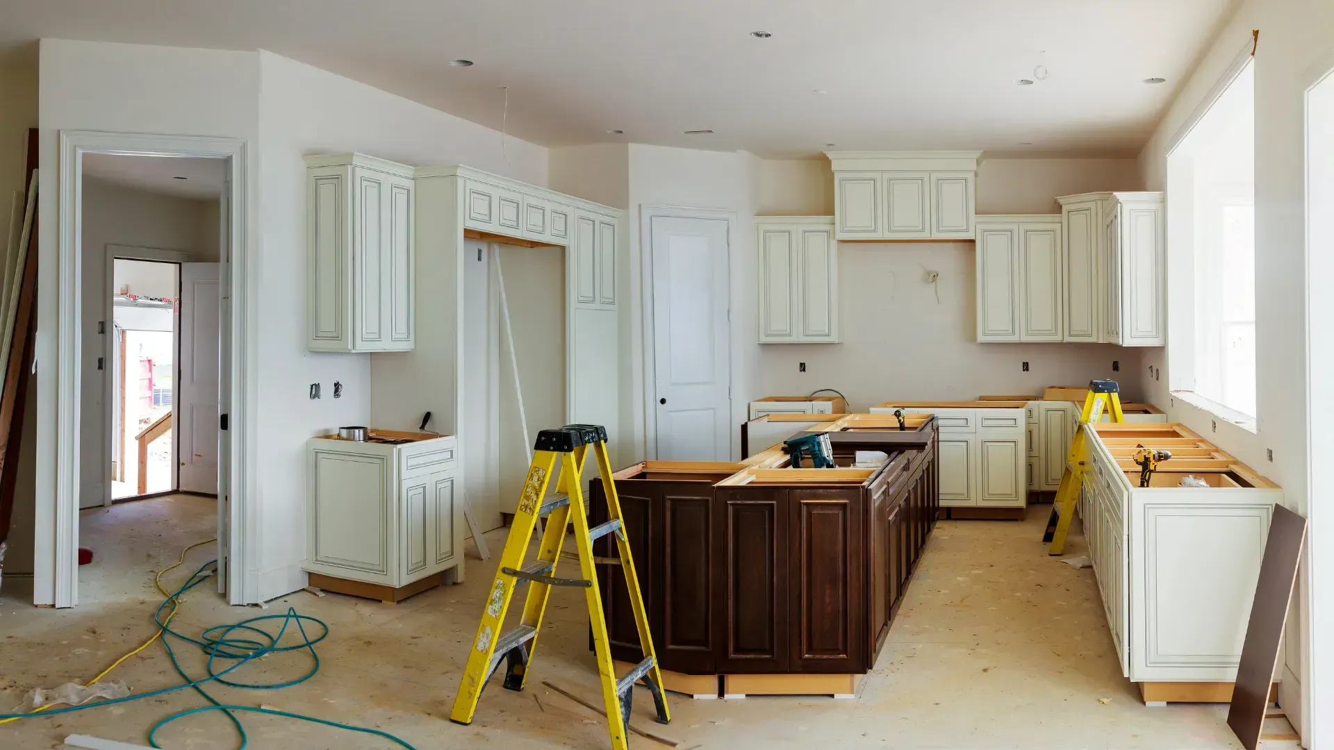 Kitchen undergoing renovation; white cabinets installed, brown island, ladder present, tools and materials visible.