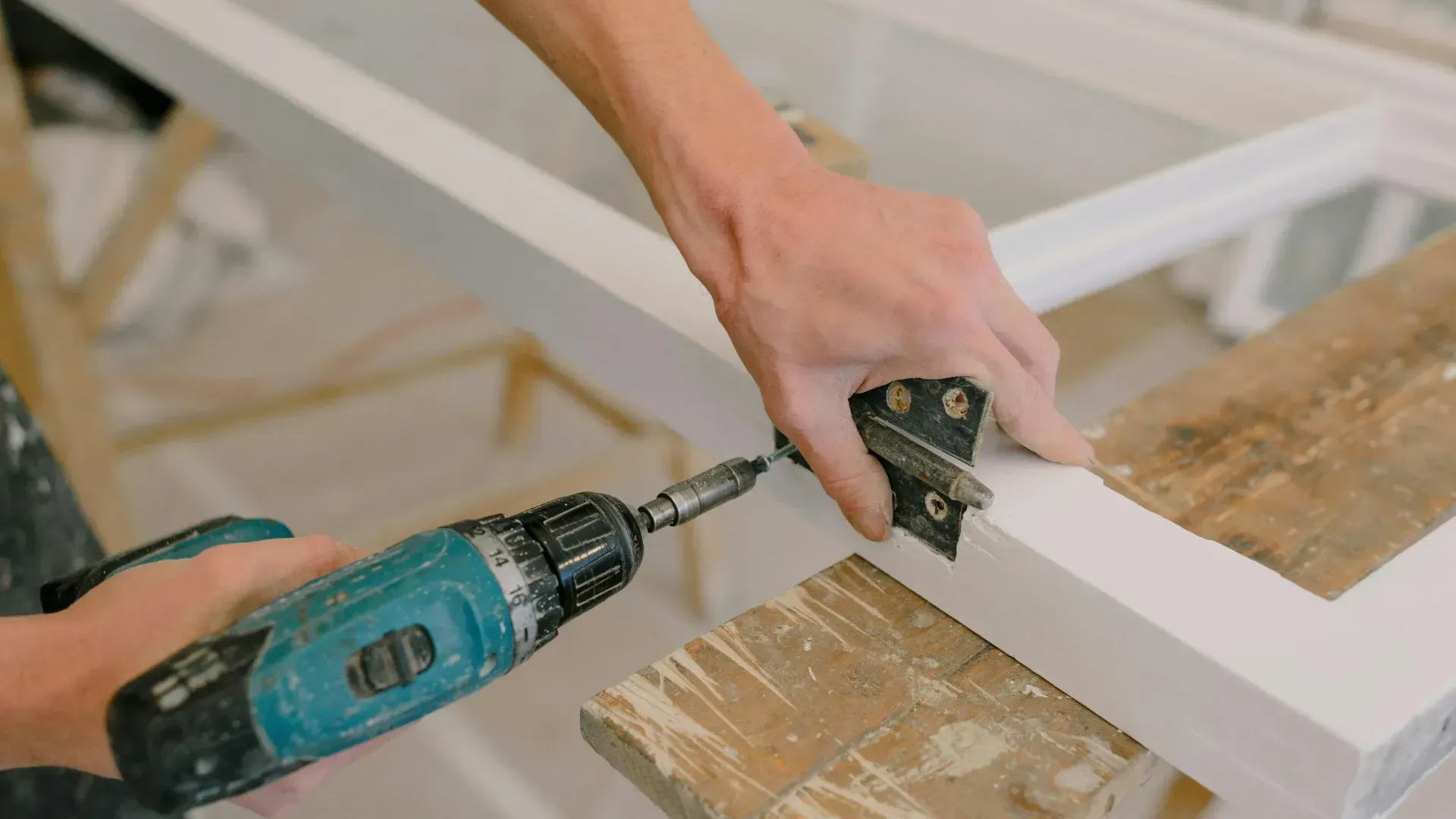Person using a power drill to attach a metal hinge to a white wooden window frame on a workbench.